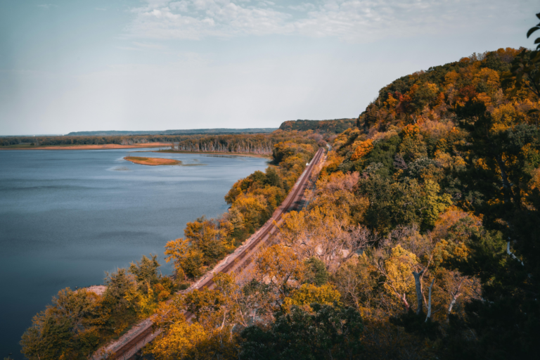A panoramic view of a railway track cutting through colorful autumn trees alongside a peaceful river and scenic bluffs, capturing the essence of interventionist in Illinois in the landscape.