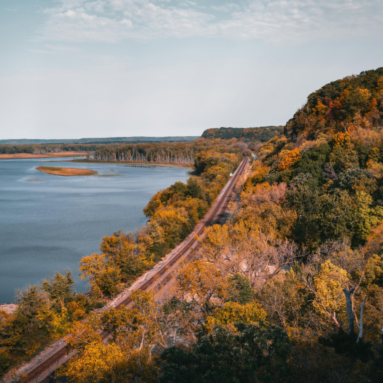 A panoramic view of a railway track cutting through colorful autumn trees alongside a peaceful river and scenic bluffs, capturing the essence of interventionist in Illinois in the landscape.